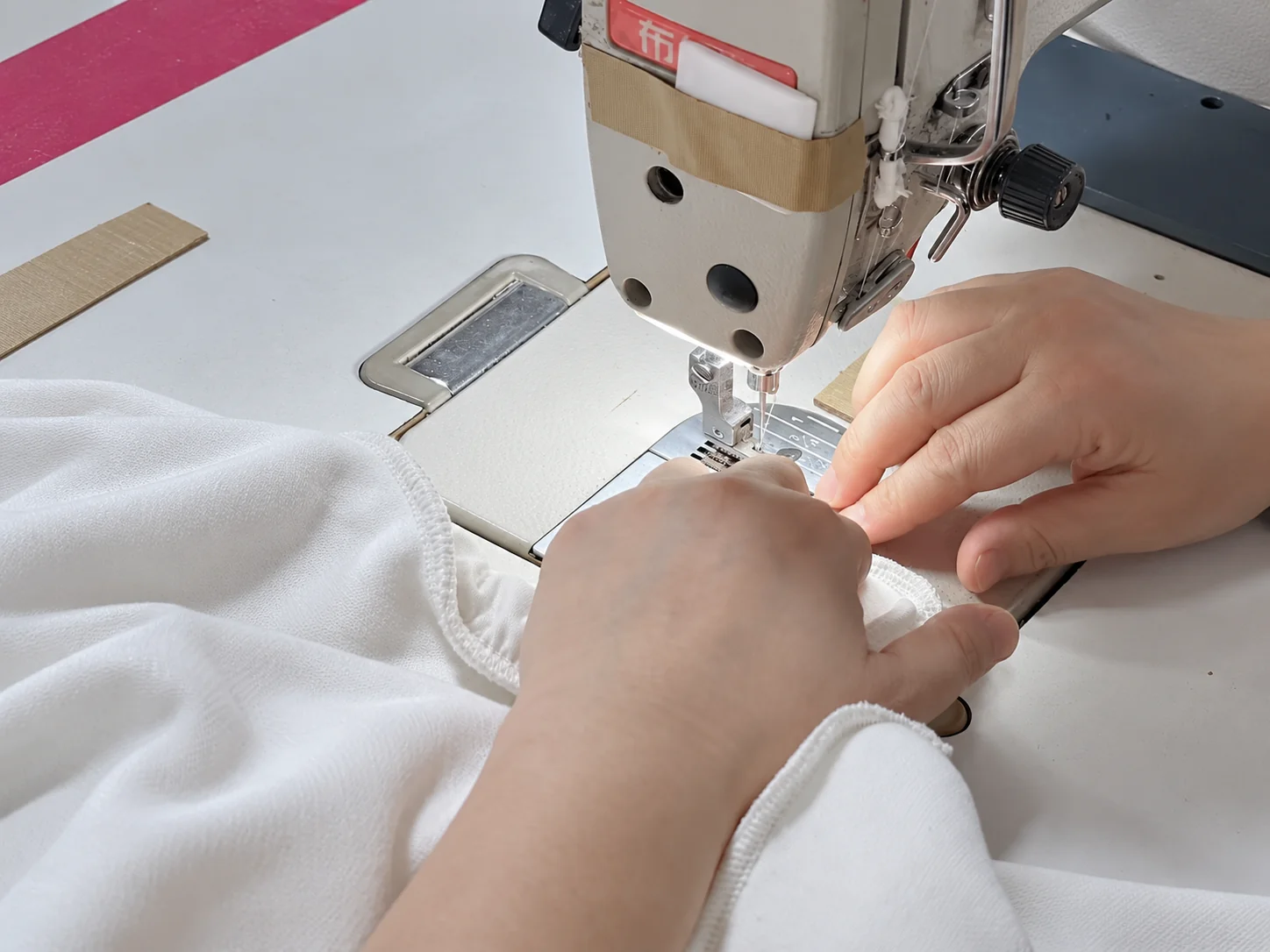 Close-up of hands guiding white garment fabric through an industrial sewing machine during sample making